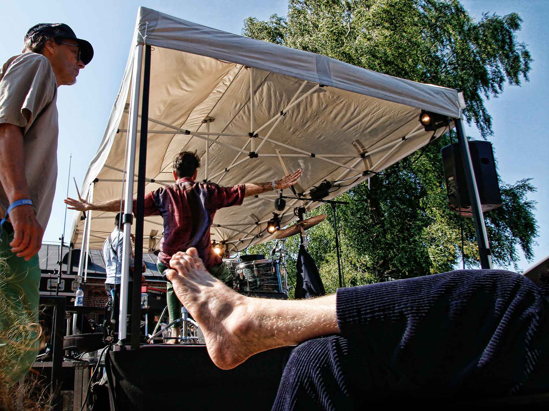 A photograph capturing a barefoot man with hairy feet reclining on the grass beside a summery stage, taken by Tony Öhberg for Finland Today.