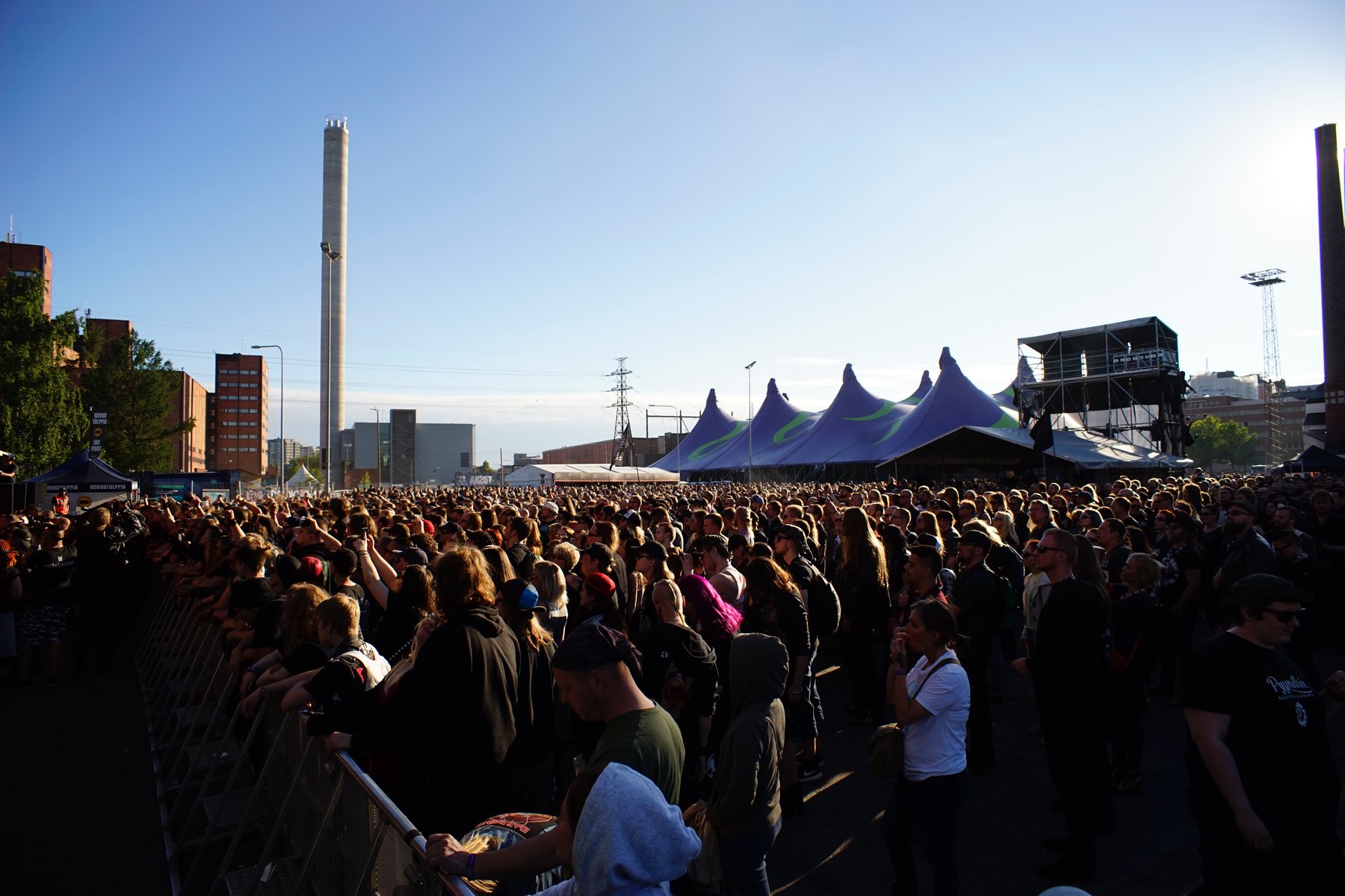 A large crowd gathers outdoors at the Tuska Festival in Suvilahti, Helsinki, with a stage and festival structures visible under a summer sky. Photograph by Morgan Walker for Finland Today.